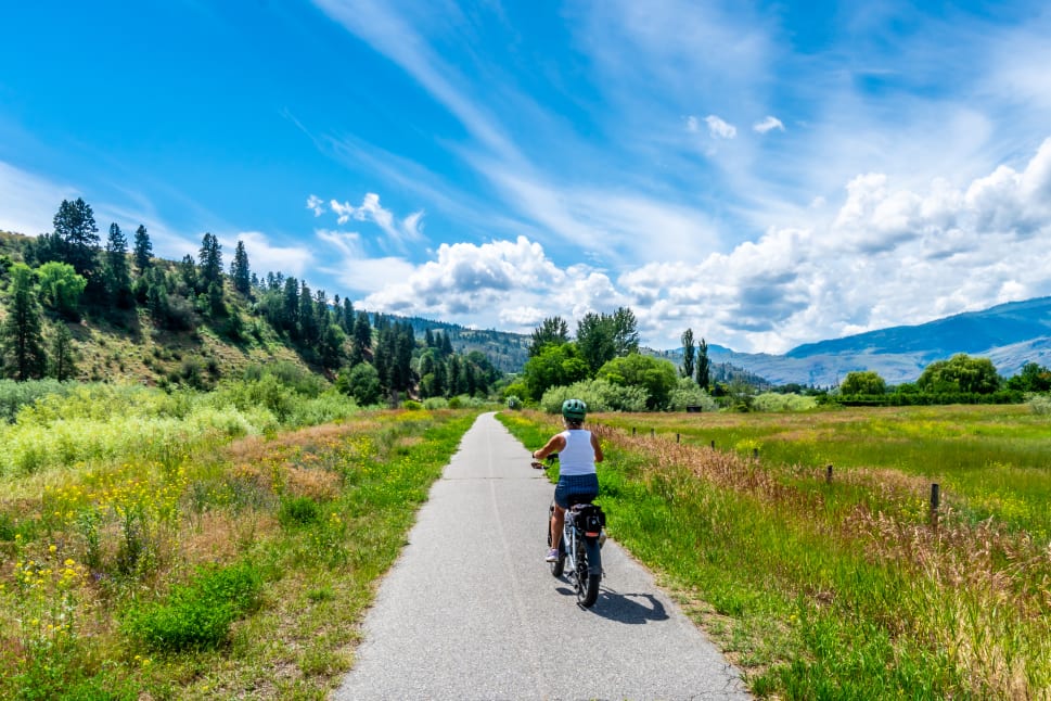 A person riding a bicycle on a paved, scenic path through the lush green landscape of Osoyoos, with hills, trees, and a vibrant blue sky with scattered clouds in the background.