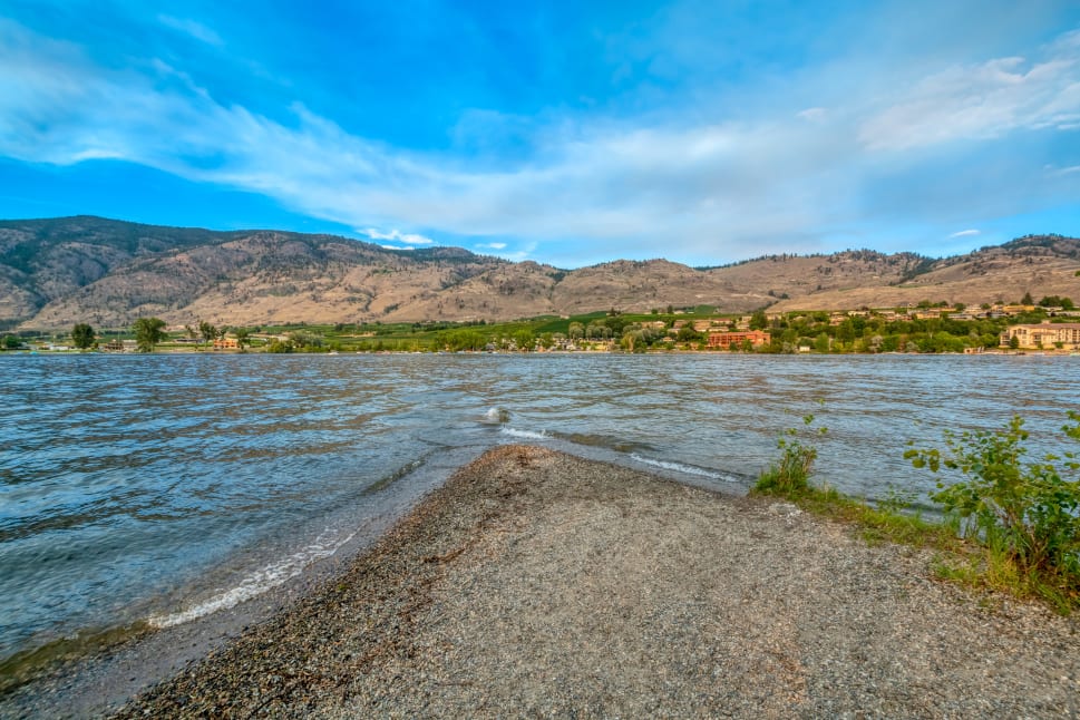 A pebbled shore leading to a calm lake with distant mountains and a partly cloudy sky in Osoyoos.