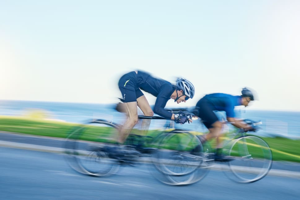 Two cyclists in motion biking along a road near the ocean with blurred background indicating speed.
