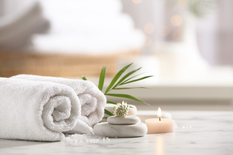 A spa setting with rolled white towels, lit candles, a palm leaf, and stacked stones on a marble surface.