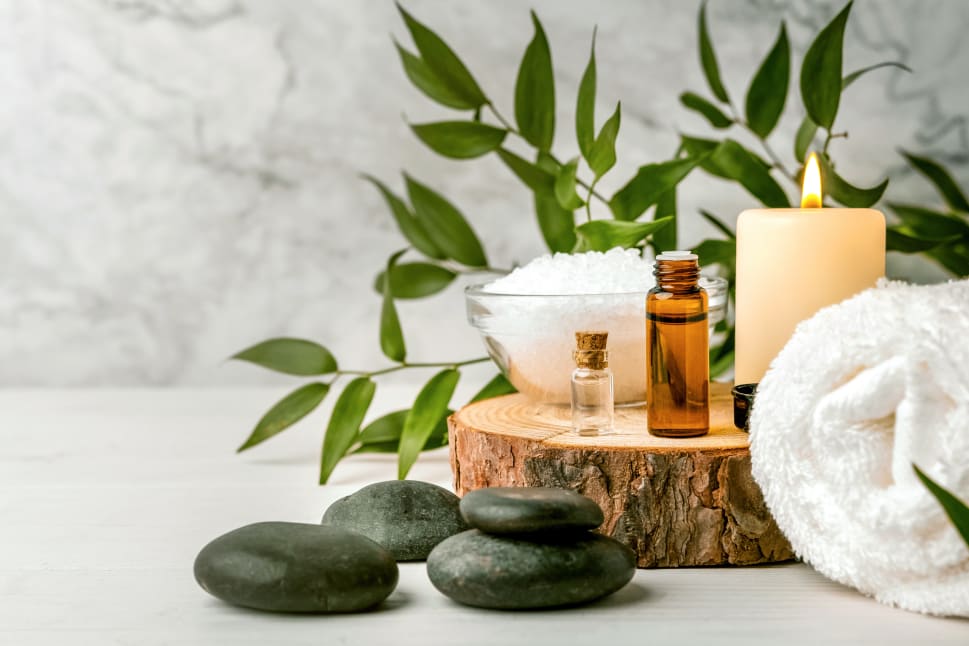 A serene spa setup with a rolled towel, burning candle, essential oil bottles, bowl of bath salts, and smooth massage stones, surrounded by green leafy branches.