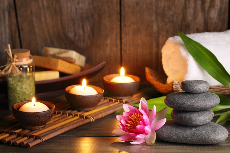 Spa scene with lit candles, stacked stones, a pink flower, soap bars, and a rolled white towel on a wooden surface.