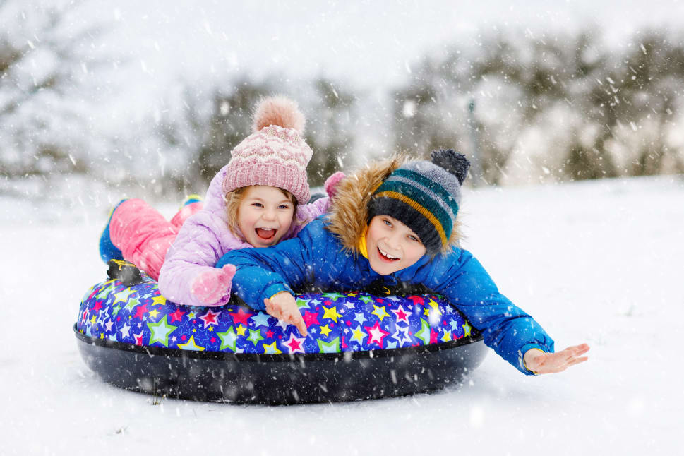 Two smiling children in winter clothes sliding down a snowy hill on a colorful inflatable tube during a snowfall.