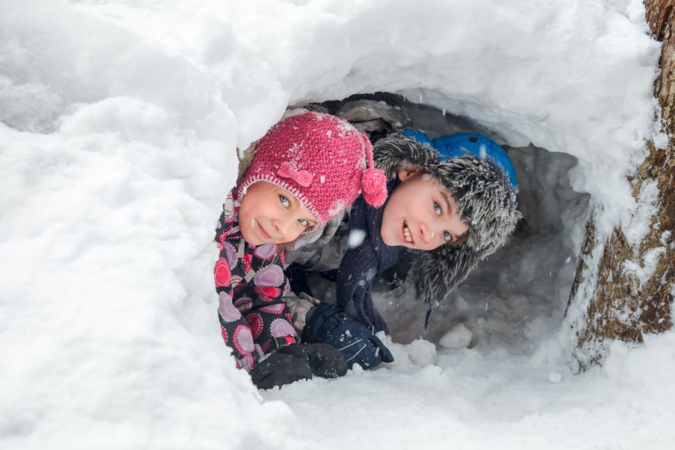 Two children, dressed in colorful winter clothing, smile while playing inside a snow tunnel during a winter activity.