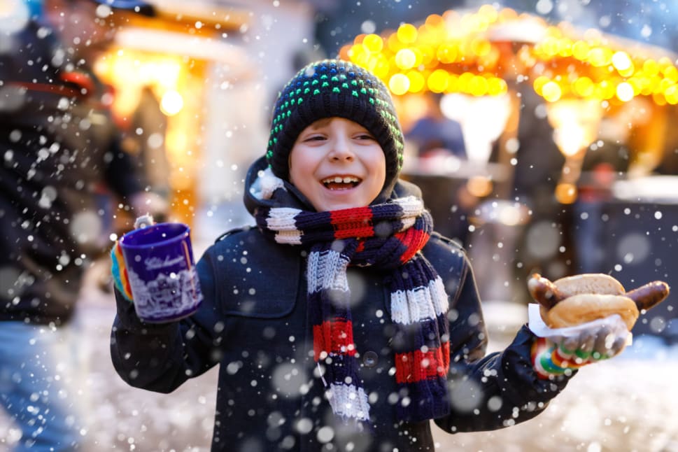 A child bundled up in winter clothes, holding a mug in one hand and food in the other, smiling joyfully in a snowy outdoor market.