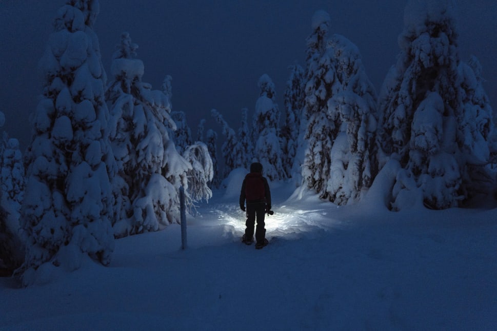A person snowshoeing in a snowy forest at dusk with a flashlight illuminating the path.