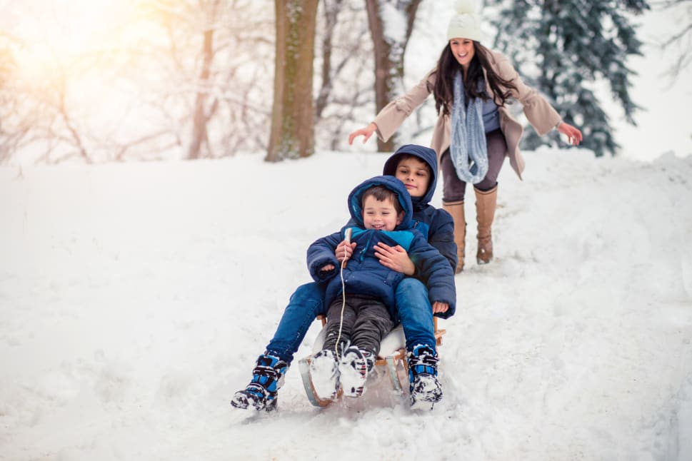 Two children on a sled sliding down a snowy hill while an adult woman pushes them from behind.