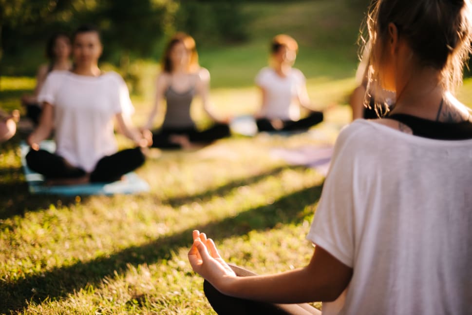 A group of women are sitting on the grass in a park, with some of them in yoga poses, and others are relaxing.