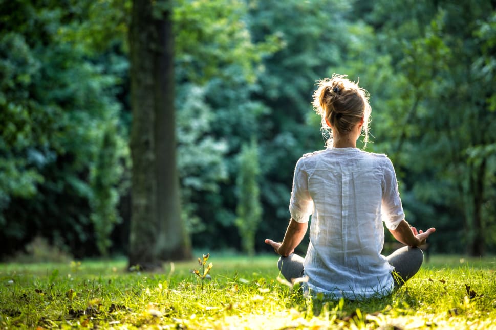A young girl is sitting in a grassy field, facing the camera, with her hands in her lap and a peaceful expression, practicing yoga.