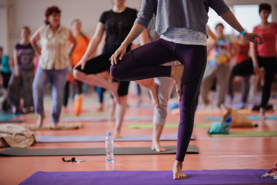 A group of people are practicing yoga in a room, with a woman in a purple leotard and a man in a gray shirt standing on their yoga mats.