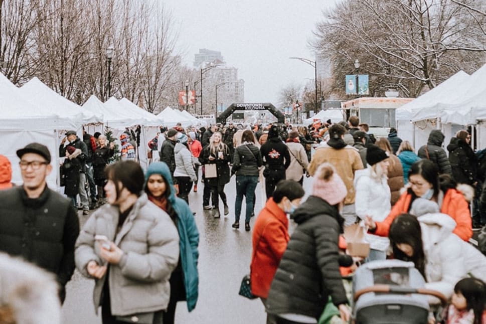 Crowds of people, bundled in winter clothing, explore the white tent booths at the Kelowna Winter Market on a wintry street in Kelowna.