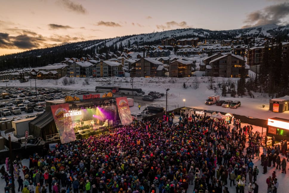 An aerial view of a large crowd gathered for a concert at Altitunes Big White with snow-covered buildings and a mountain backdrop at sunset in Kelowna.