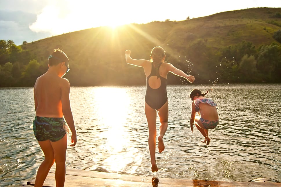 Three kids enjoying lake activities, with one jumping into the water, another mid-air, and a third standing on the dock, under the bright sun with a hilly background.
