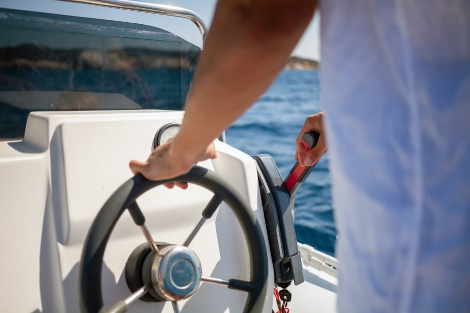 A person steering a boat on a lake, with one hand on the wheel and the other on the throttle lever.