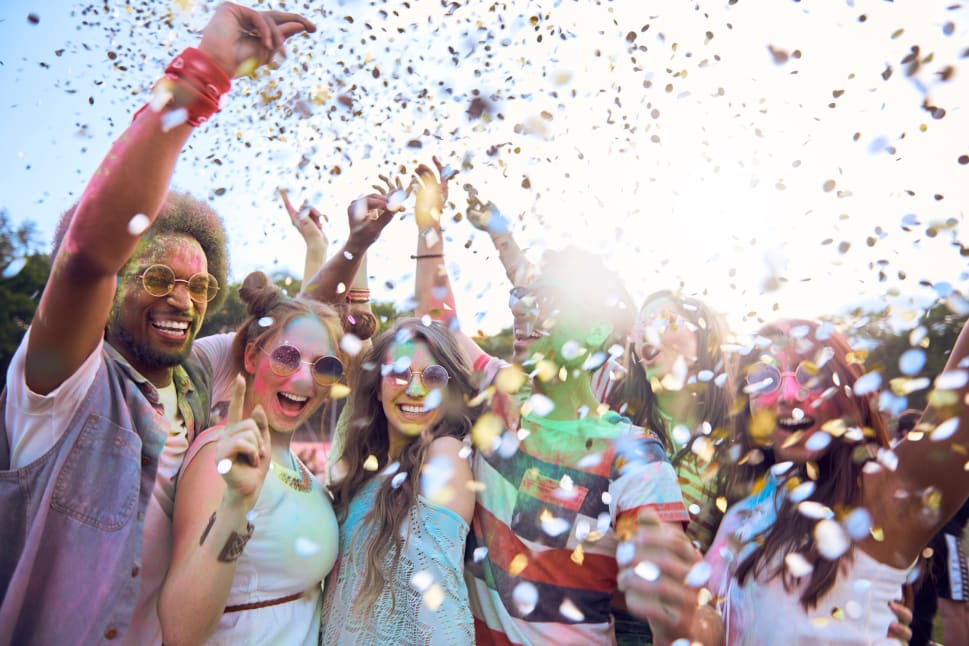 A group of friends celebrates a colorful festival with confetti flying in the air while smiling and laughing.
