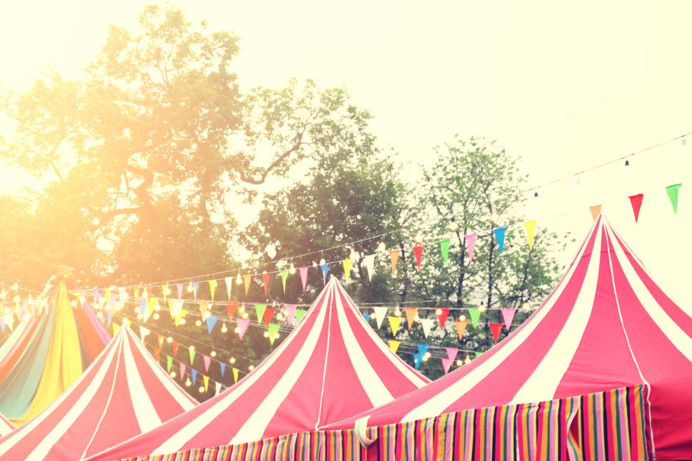 A festival scene with colorful striped tents and strings of multi-colored flags and lights in an outdoor setting with trees in the background.