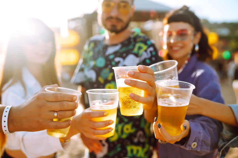 A group of people at a festival raising plastic cups filled with beer in a celebratory toast under sunlight.