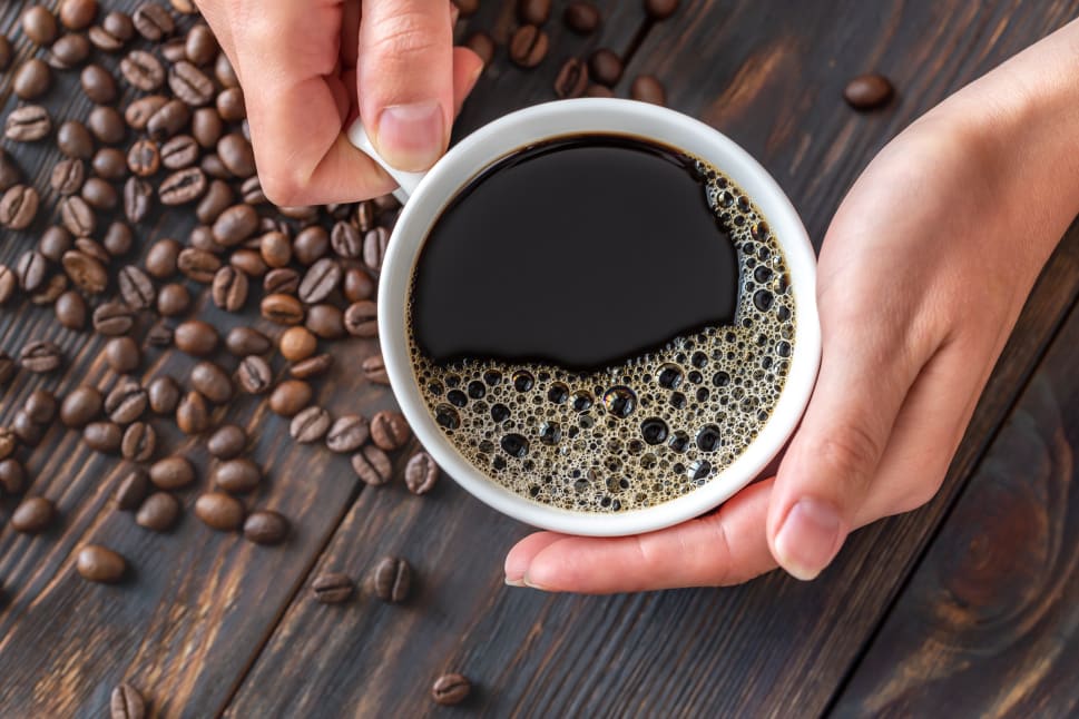 Hands holding a cup of coffee with coffee beans scattered on a wooden surface.