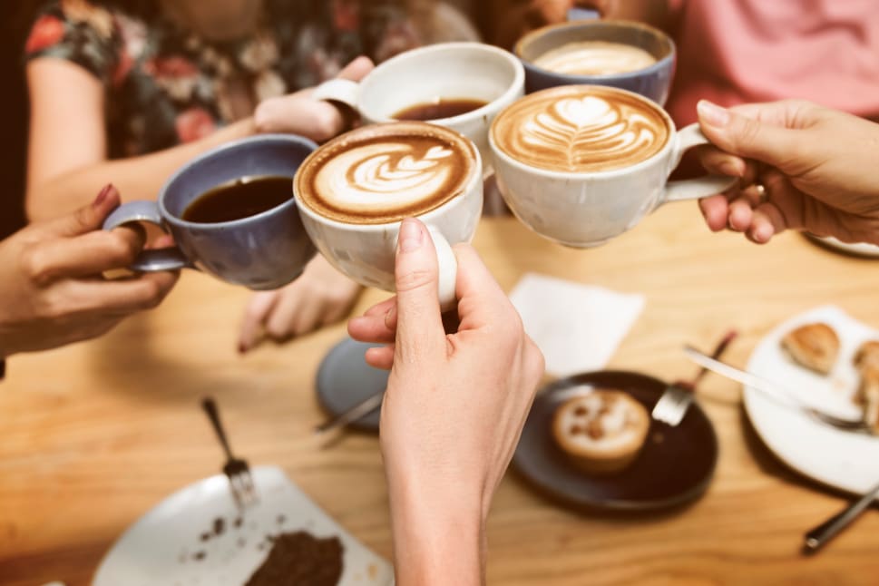A group of people toasting with coffee mugs above a table with food and pastries.