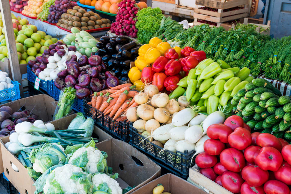Fresh vegetables and fruits including tomatoes, carrots, bell peppers, eggplants, and greens displayed at a farmers market stall.