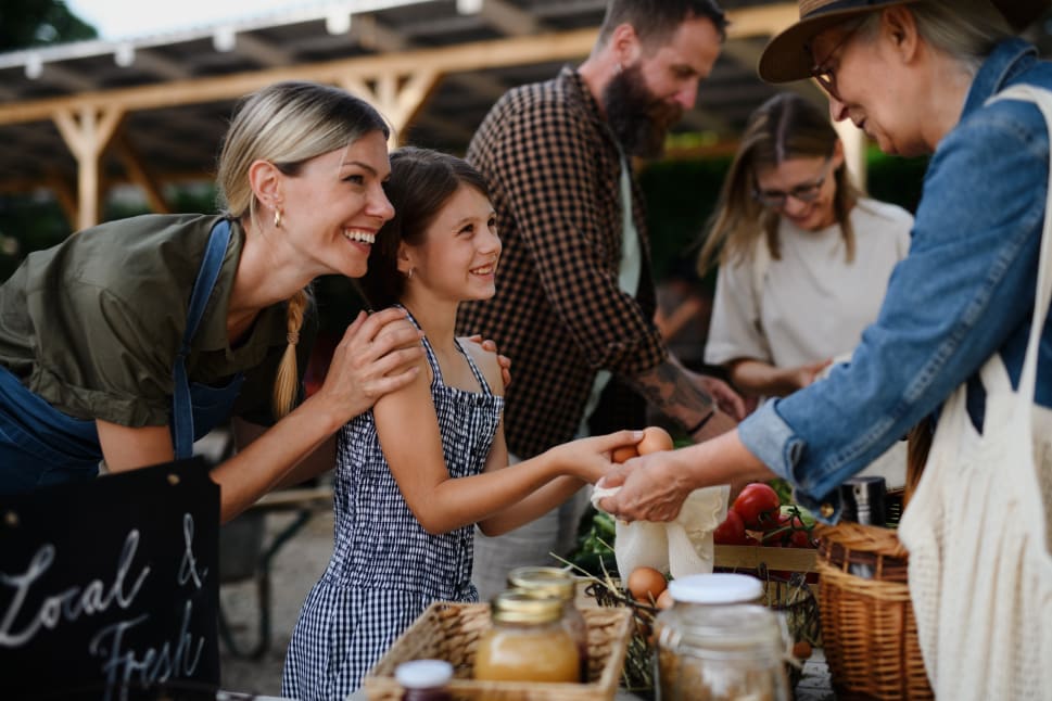 A smiling woman and her daughter buy fresh produce from an elderly vendor at a bustling farmers market.