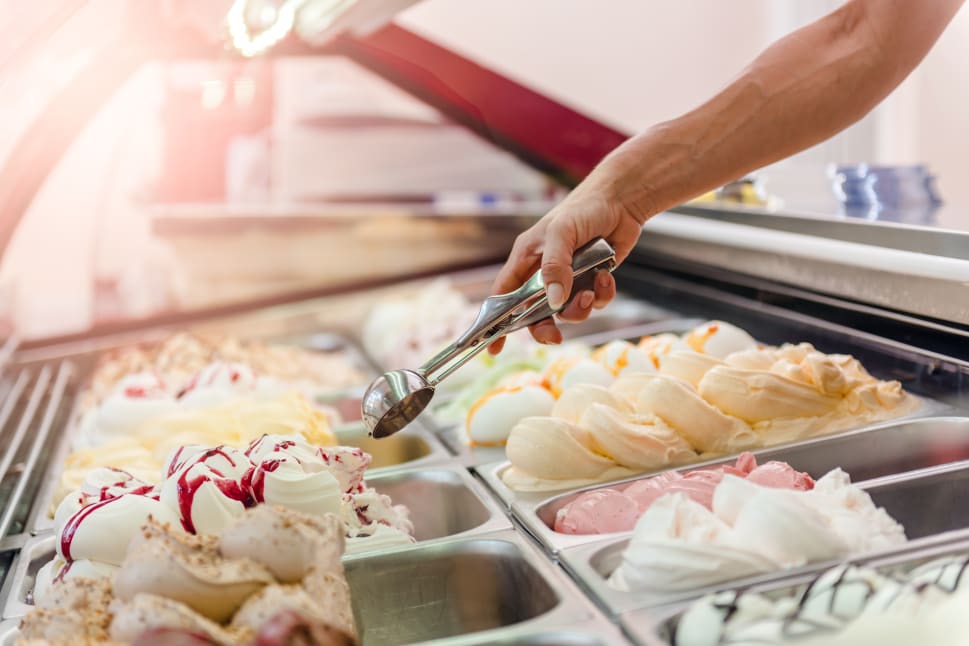 A hand holding a scoop over a display of various flavors of ice cream or gelato in a store.