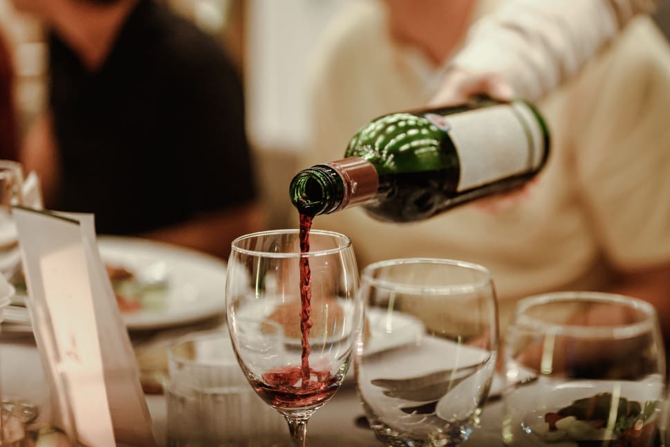 A green wine bottle is being poured into a glass at a dinner table surrounded by blurred people and tableware.