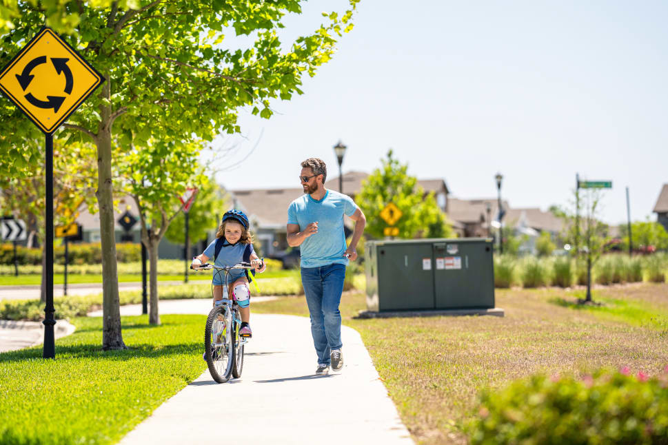 A father jogs alongside his young daughter as she rides her bike on a sunny neighborhood path with trees and a roundabout sign nearby.