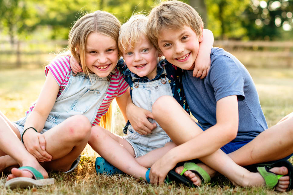 Three smiling children sit close together on the grass in a park, hugging each other and enjoying a sunny day.