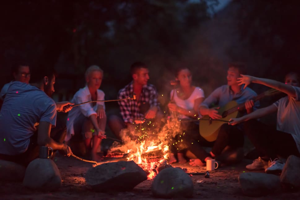 A group of people sitting around a campfire at night, with one playing a guitar and others roasting marshmallows on sticks.