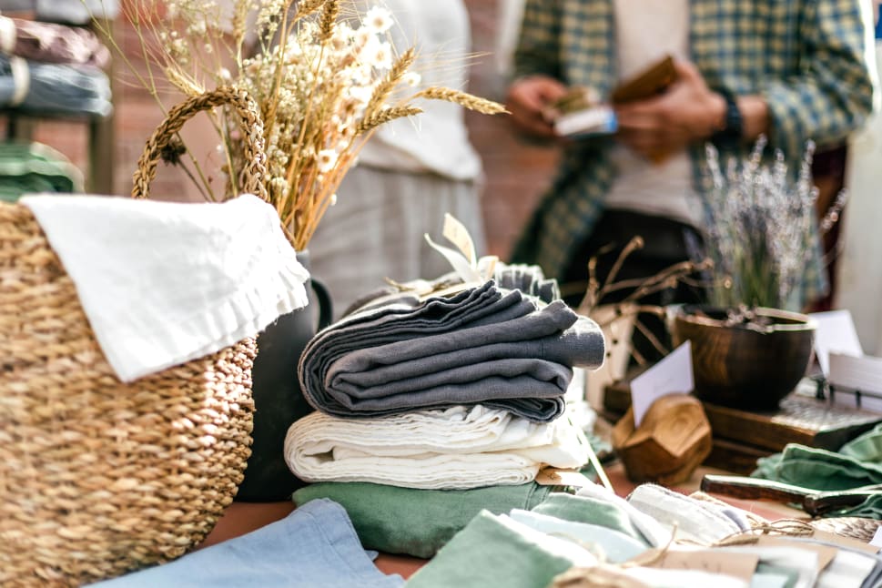 A display of folded fabrics, woven basket with dried flowers, and various home decor items on a table at a shopping market.