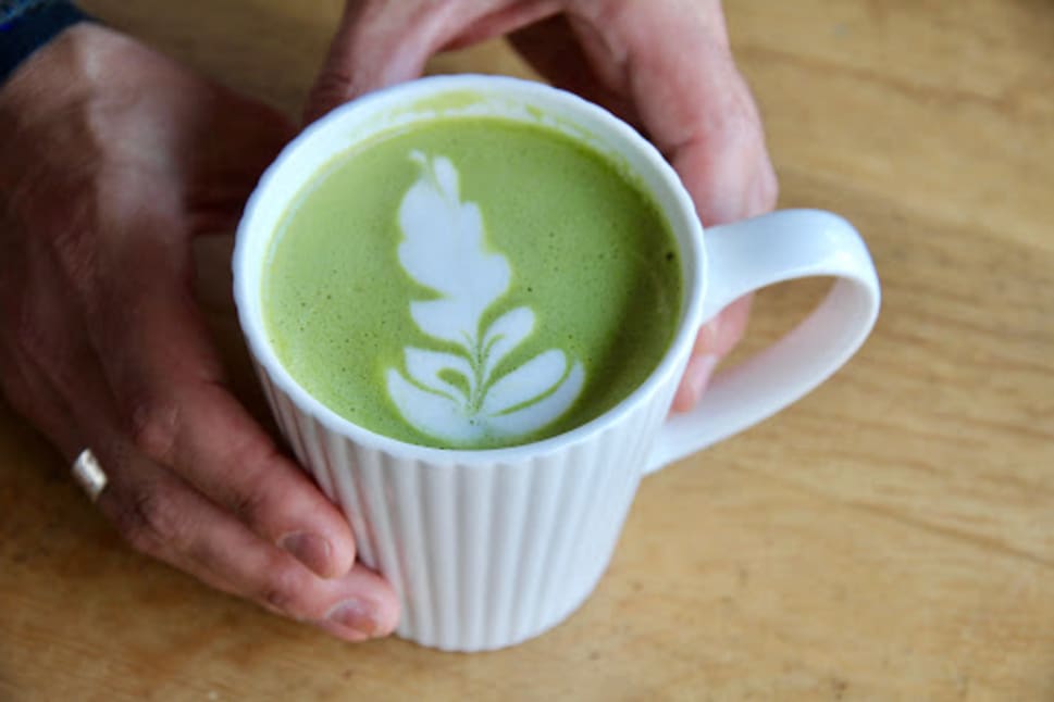 A person's hands holding a white cup filled with matcha, decorated with a leaf latte art, placed on a wooden table.
