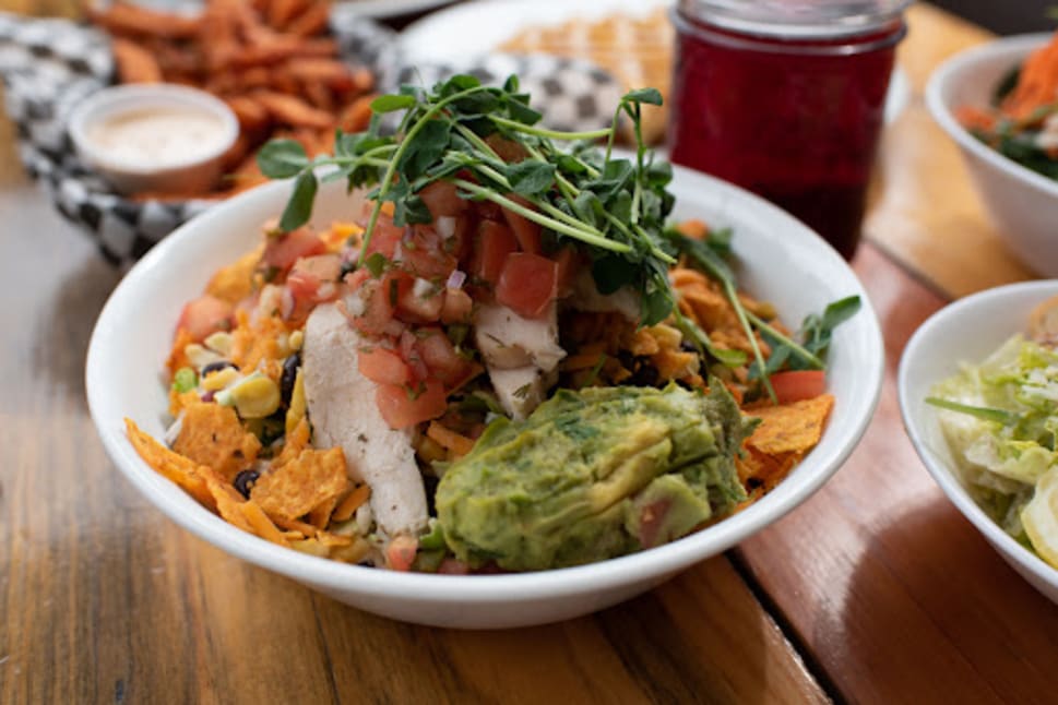 Plate of vibrant, health-conscious mixed salad with chicken, guacamole, and fresh vegetables on a wooden table at Eatology restaurant in Vernon, BC.