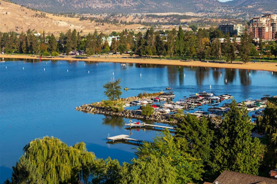 A picturesque view of a serene lake with a dock and boats, surrounded by lush green trees and mountains in the background at Kettle Valley Beach Resort in Penticton.