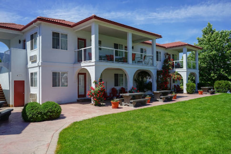 Resort-style building with white exterior, red roof, balconies, lush garden, and outdoor seating area in Sandy-Beach-Suites, Osoyoos.