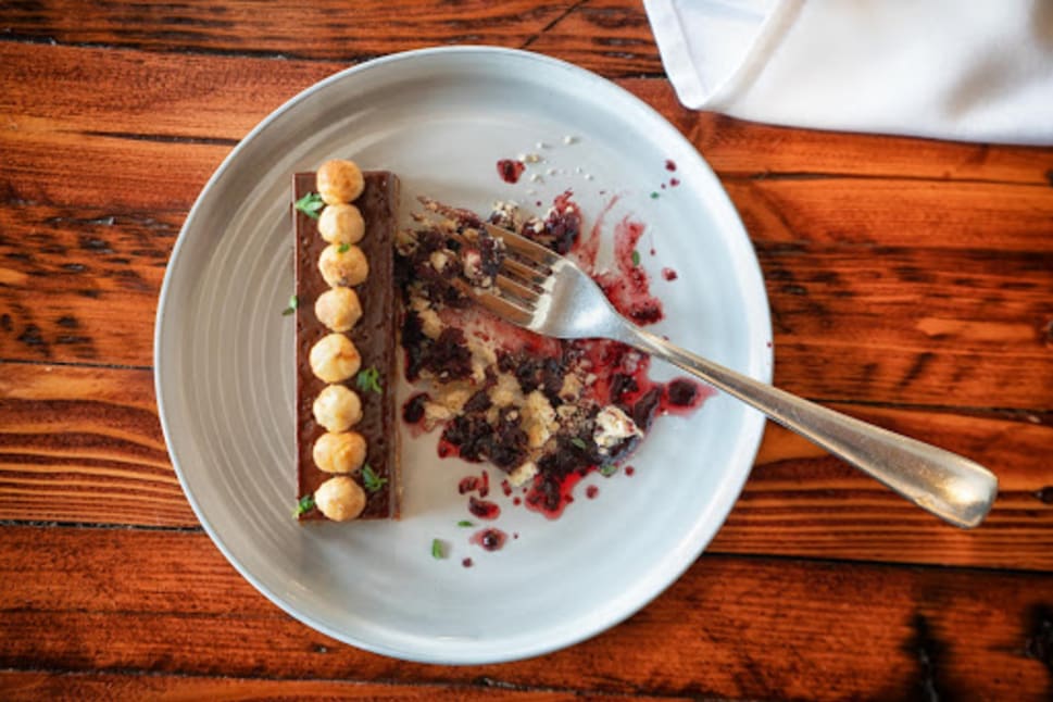 A gourmet dessert with a chocolate base topped with round confections on a white plate, served with a fork on a wooden table.