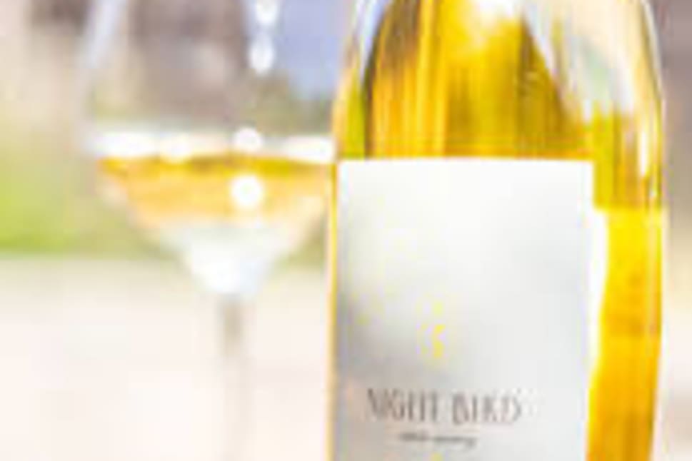 A bottle of white wine labeled "Night Bird Estate Winery" with a filled wine glass blurred in the background.