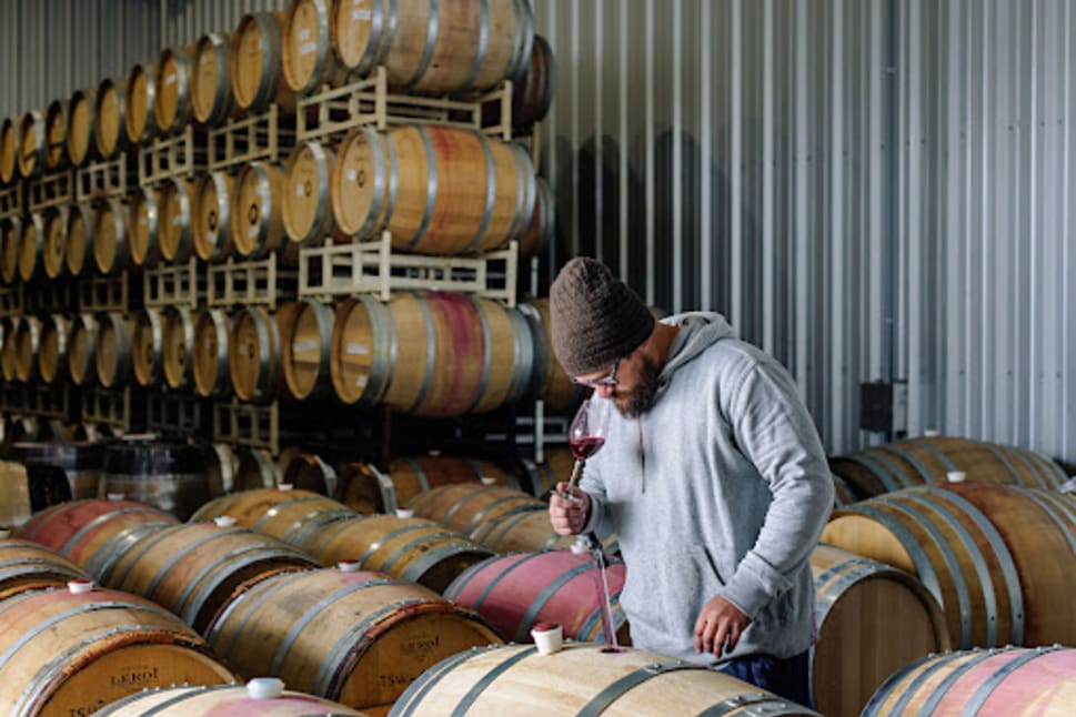 A person at Three Sisters Winery in Penticton, BC, tasting wine from a barrel in a wine cellar filled with stacked barrels.
