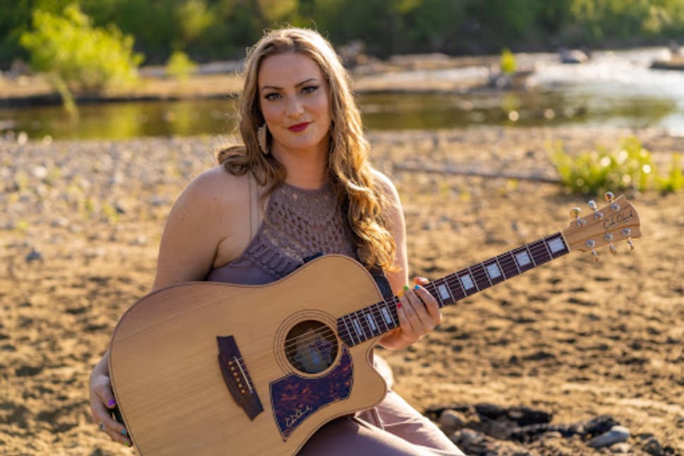 A woman in a sleeveless top sits on the riverbank playing an acoustic guitar, with greenery and water in the background under natural sunlight.