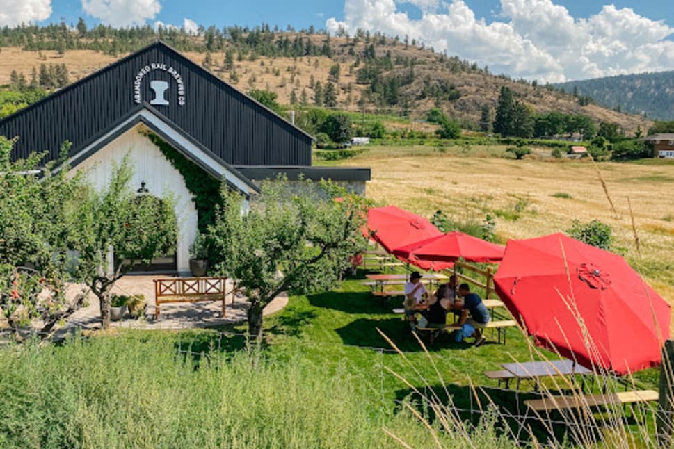A scenic view of Abandoned Rail Brewing Co. in Penticton with outdoor seating under red umbrellas and a picturesque hillside in the background.