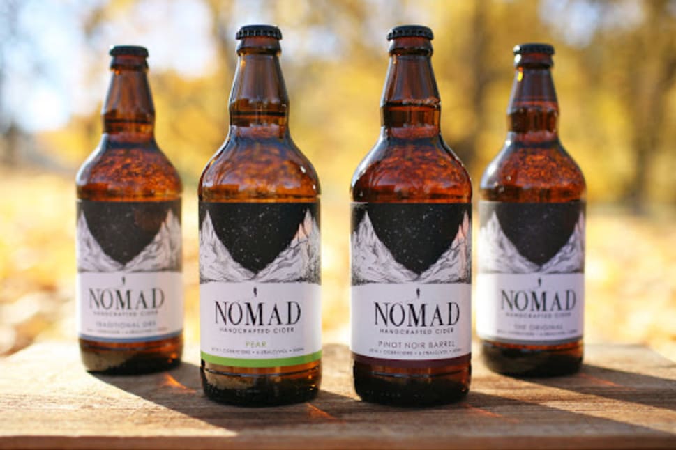 Four bottles of NOMAD handcrafted cider placed on a wooden surface with an outdoor background showcasing autumn colors.
