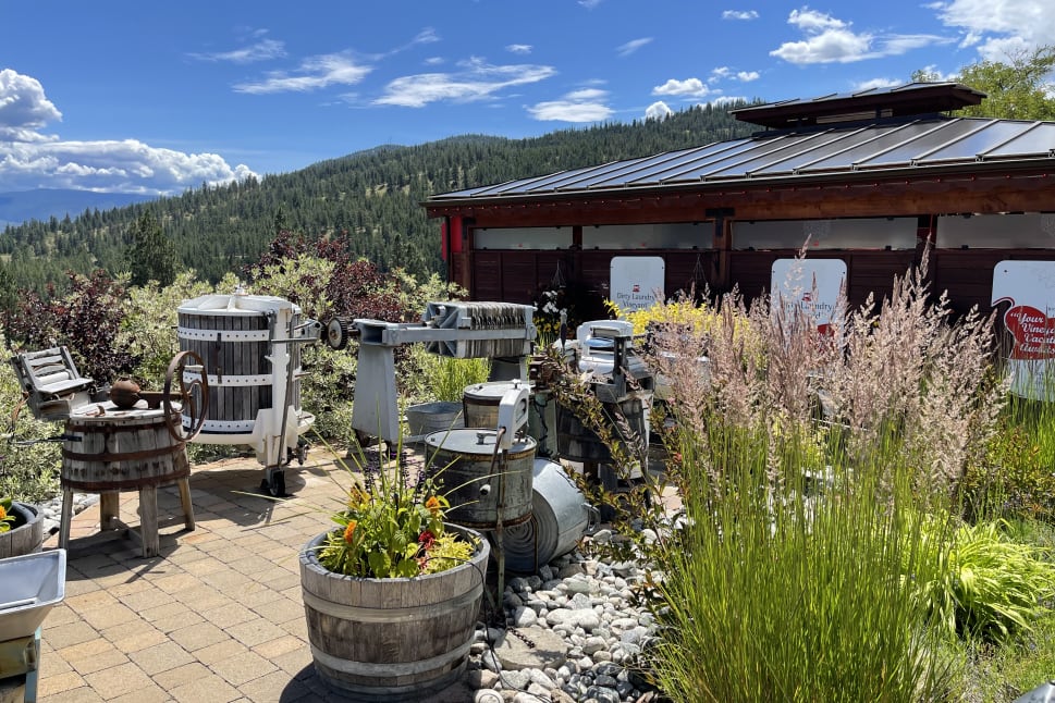 Vintage winemaking equipment set in a scenic outdoor courtyard with a view of lush hills under a blue sky in Summerland.