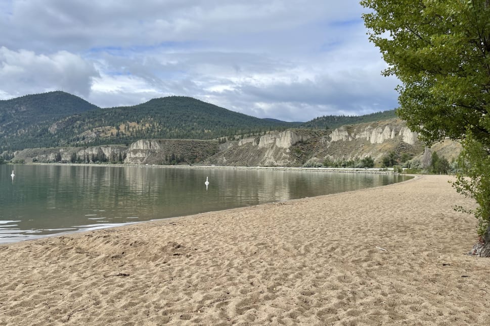 Scenic view of a sandy beach leading to a calm lake surrounded by wooded hills and cliff formations under a partly cloudy sky in Summerland.