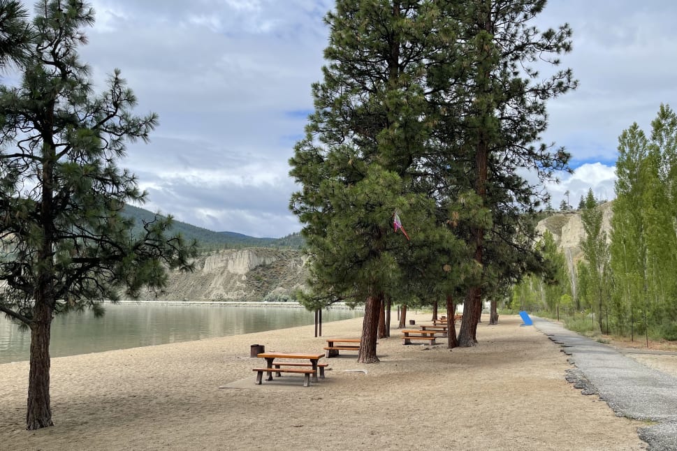 A peaceful lakeside beach at Summerland, with picnic tables and tall trees under a cloudy sky, mountains in the background.