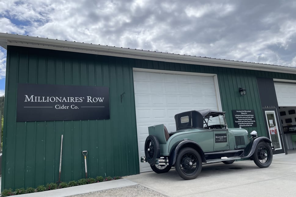 Green vintage car parked outside Millionaires' Row Cider Co. building under a cloudy sky in Summerland.