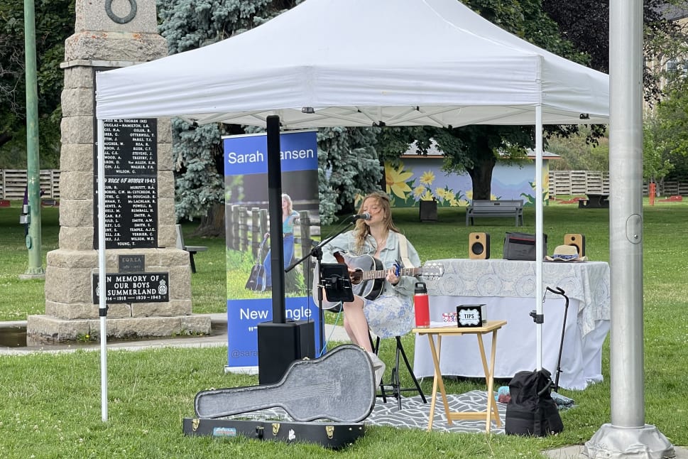A singer performs with a guitar under a white tent in a park in Summerland, near a memorial, with promotional signage and audio equipment set up.