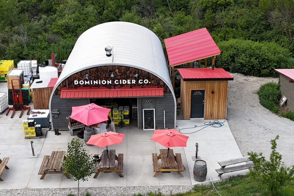 Outdoor view of Dominion Cider Co. in Summerland with picnic tables, red umbrellas, and equipment storage surrounded by greenery.