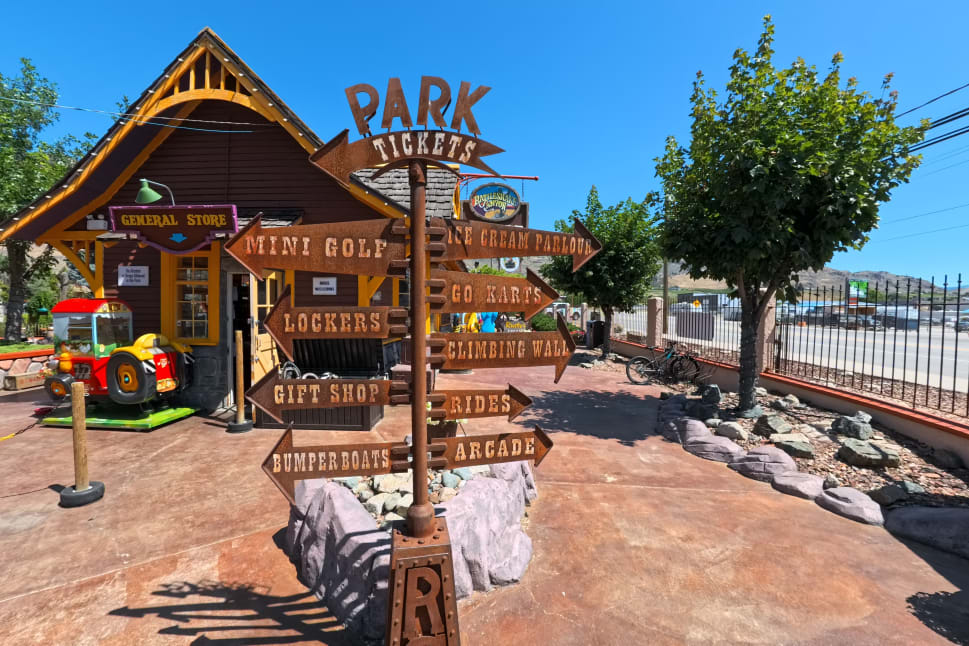 A park signpost in Osoyoos directing to various activities like mini golf, ice cream parlour, go-karts, and rides, in front of a general store.