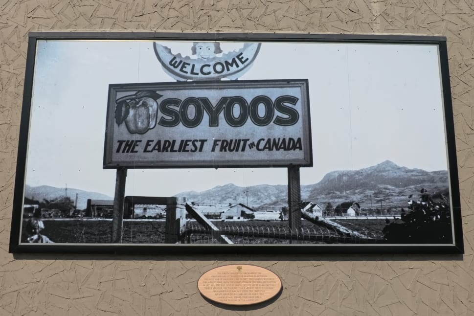 A vintage sign reads "Welcome to Osoyoos: The earliest fruit in Canada" with a scenic backdrop of mountains and buildings.