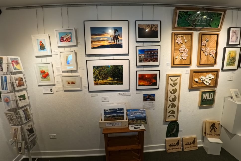 An art gallery in Osoyoos displaying various framed paintings, photographs, and woodwork on white walls, with a rack of greeting cards on the left.